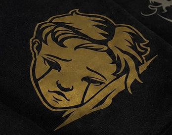 A folded black t-shirt displayed on a dark wooden surface, featuring gold and silver graphic prints including a stylized face, a snake, and a shield emblem with the word 'OBLIVION'.